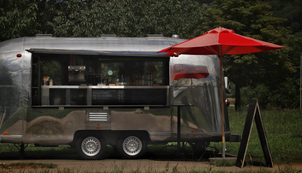 A chrome food trailer with a chalkboard menu and a standing red umbrella next to the ordering window.