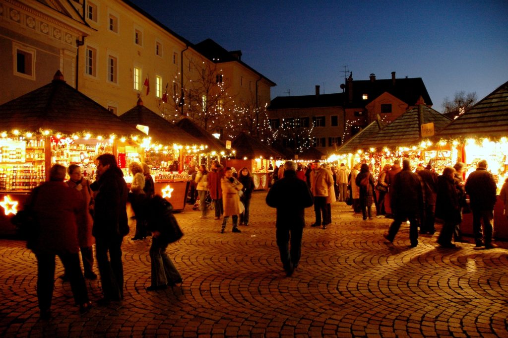 Crowds of people walk between stalls at an outdoor winter market with string lights illuminating each stall.