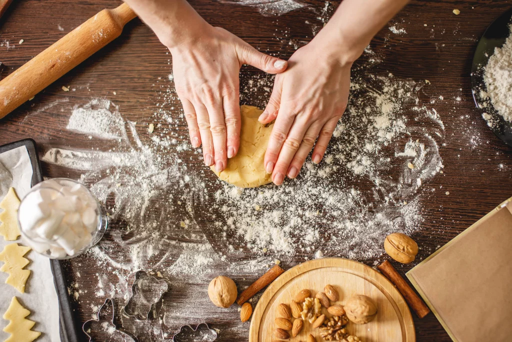 An overhead shot of a person shaping dough on a wooden table covered with flour, rolling pins, nuts, spices, and other baking items.