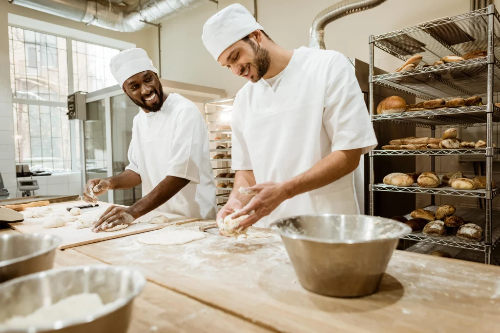 Two bakers kneading dough on a a wooden countertop in a commercial kitchen surrounded by racks of freshly baked bread.
