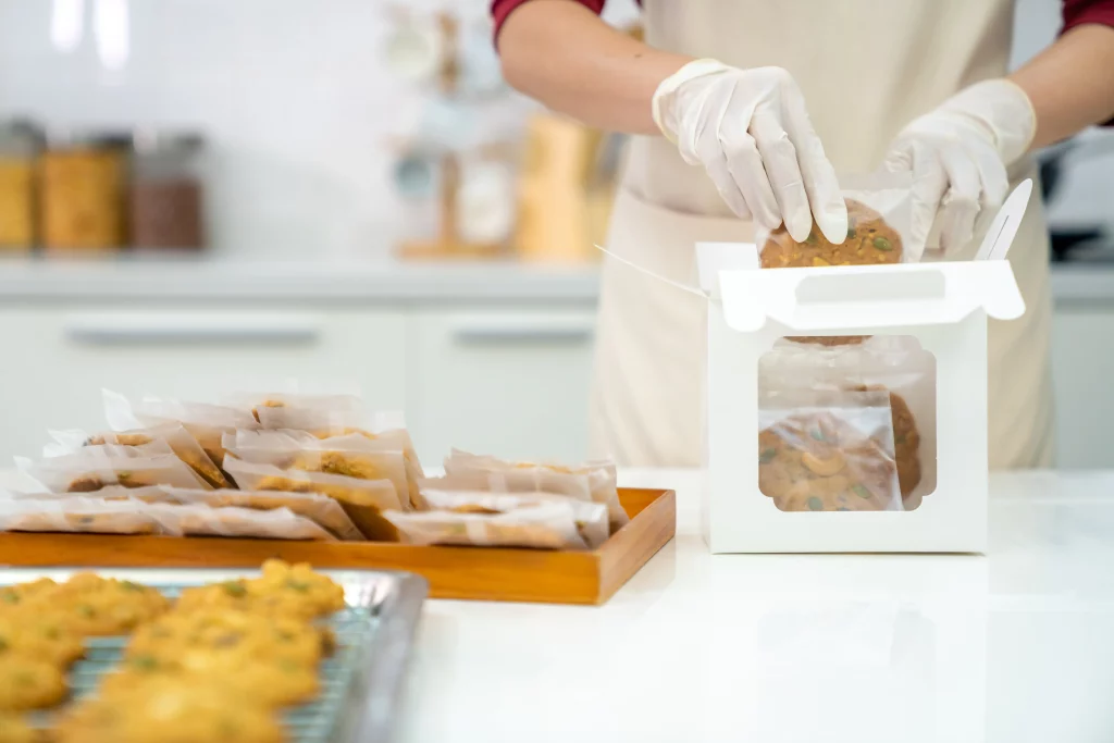 A home baker puts individually wrapped cookies into a white delivery box while wearing rubber gloves.