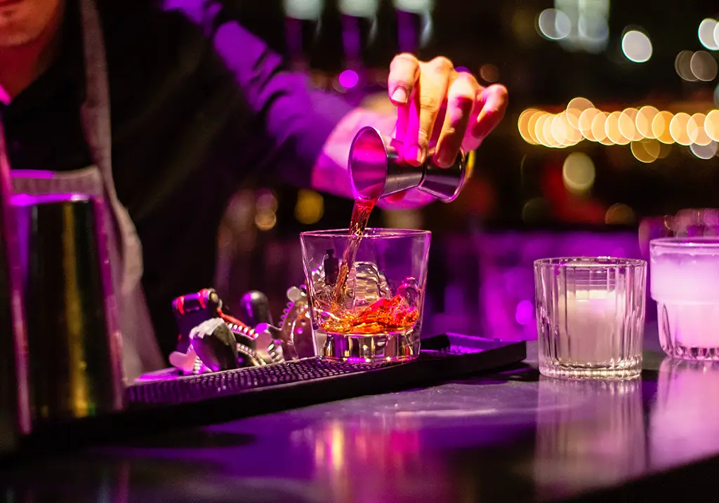 Close-up shot of a bartender pouring a shot of liquor into a glass filled with ice cubes. The bar is illuminated by purple lighting.