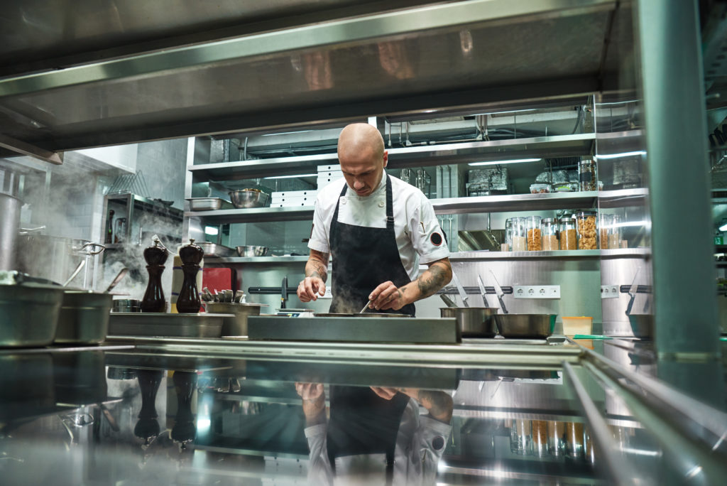 A chef prepares food in a commercial kitchen surrounded by stainless steel shelving and appliances.