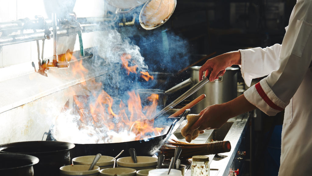 A professional chef stirring food in a frying pan with flames and steam rising out of it.