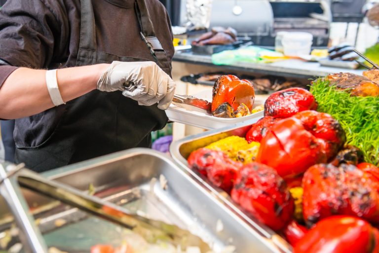 A food service worker using tongs to place a grilled red bell pepper inside a takeaway container.