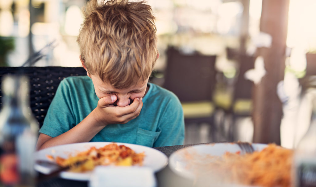 A young boy has his hand over his mouth while sitting at a table in a restaurant because he is feeling ill from the food. Product liability insurance may help in instances of illness from food products you help produce.