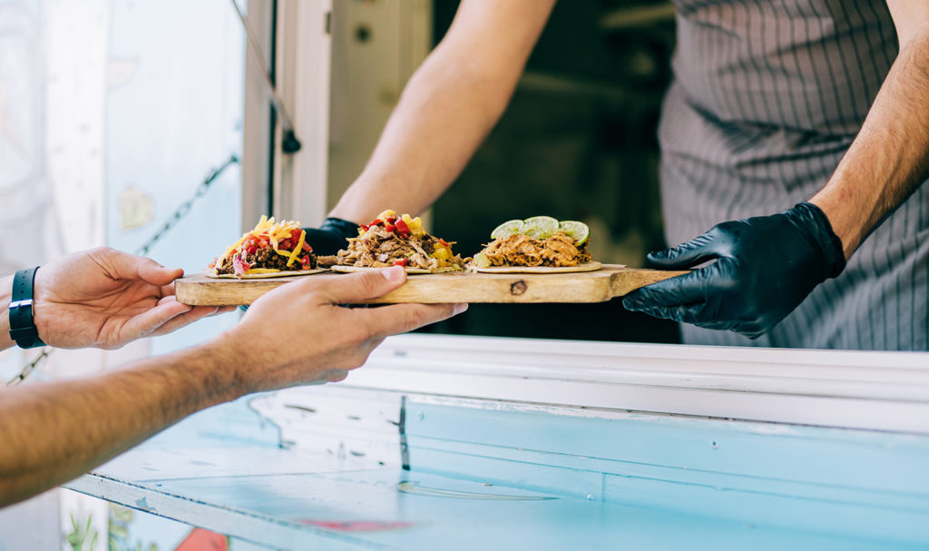 The gloved hands of a food truck chef are passing a tray of freshly made tacos out to a customer's outstretched hands. If the customer is injured or falls ill from the food, the costs may be covered with product liability insurance.