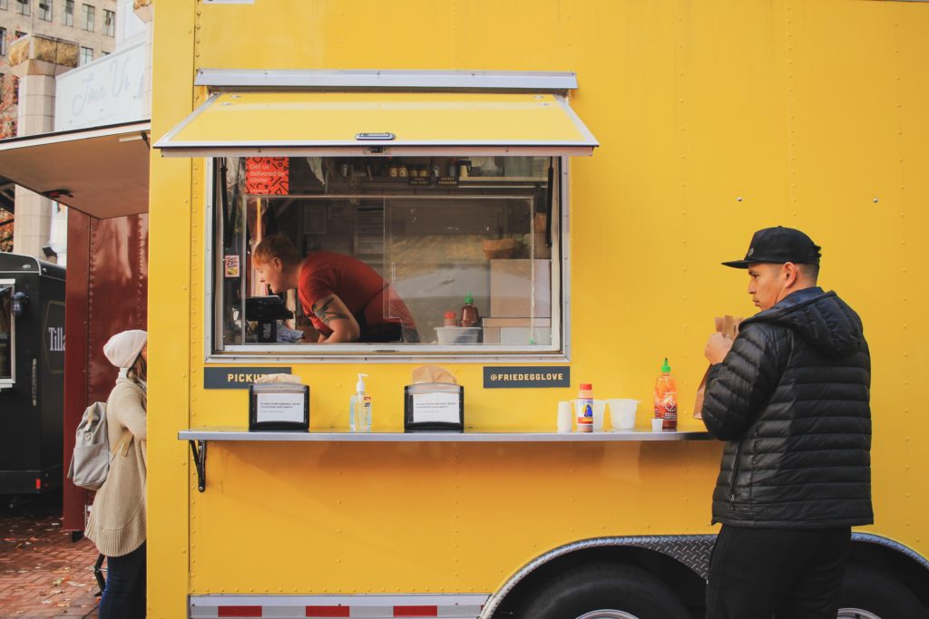 A man holds a bag of food next to the pickup window of a yellow food truck while an employee helps the next customer in line.