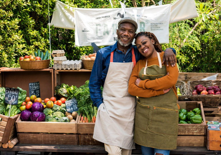 A happy couple standing in front of their produce stand at a local farmers market.