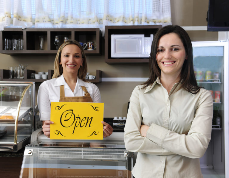 A restaurant owner smiles and poses with an employee, who is holding a sign that reads “open.”