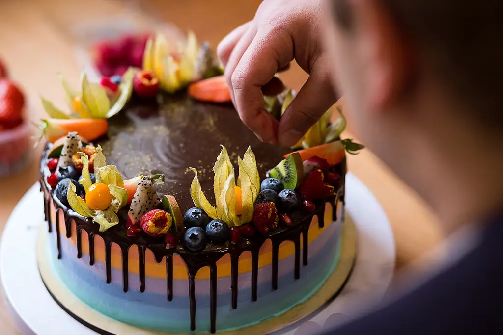 A person puts slices of kiwi and dragonfruit on the top of a colorful cake with a chocolate glaze on top.