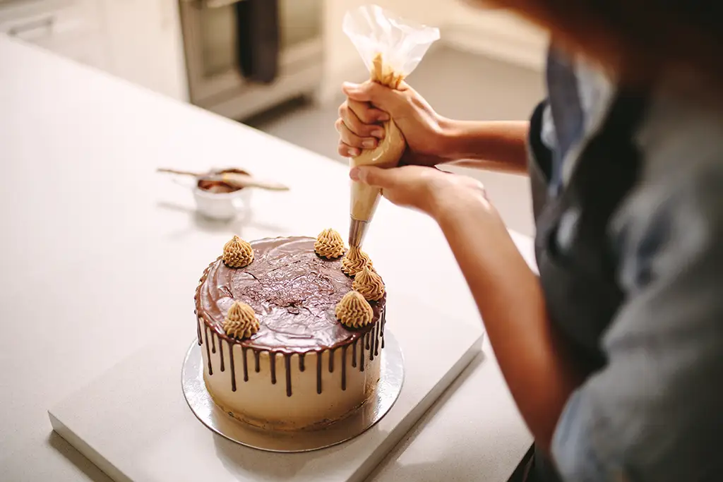 A baker uses a piping bag to put dollops of frosting on top of a cake.