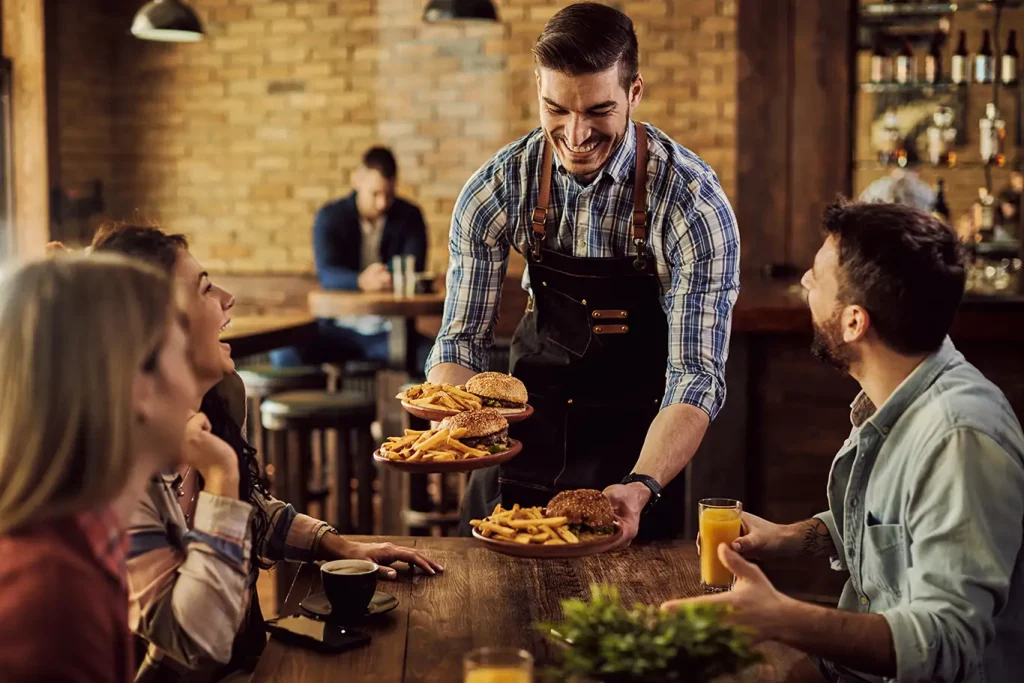 A smiling waiter serves burgers to a table of three people in a restaurant.