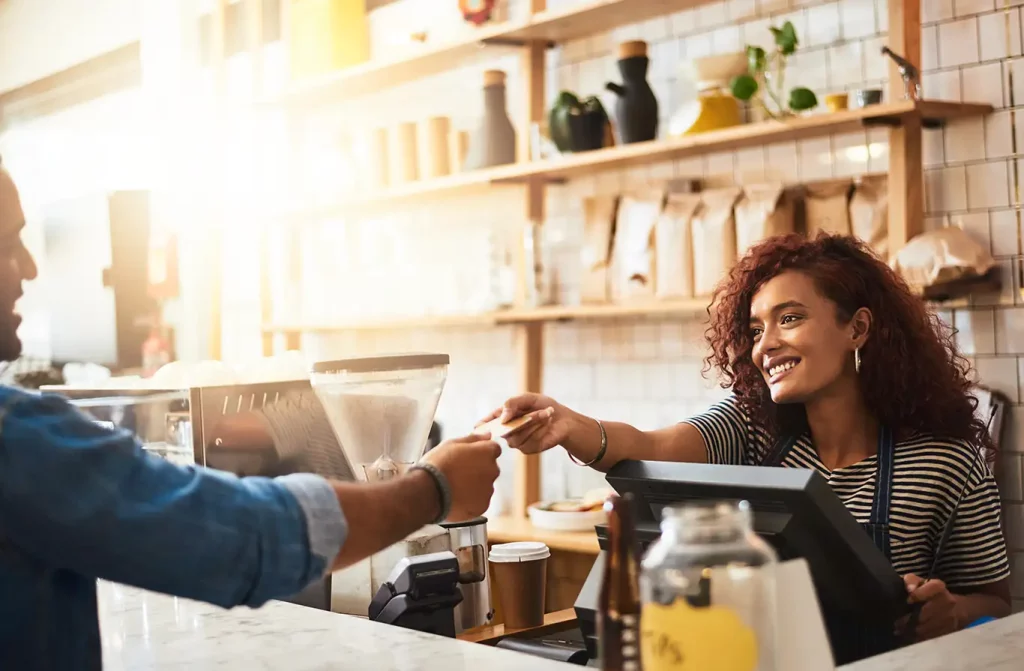 A customer at a cafe hands his credit card to a smiling cashier in a striped shirt.