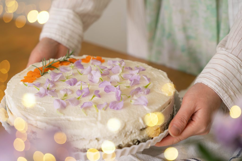 A person holds a frosted cake on a plate with purple and orange floral decorations on top.