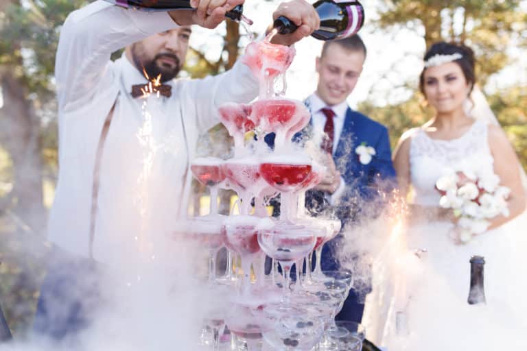 A wedding bartender pours sparkling wine down a pyramid of glasses in front of a bride and groom.