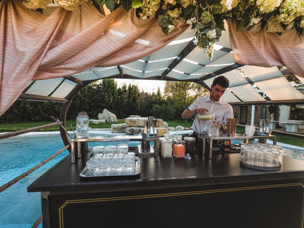 A bartender prepares drinks at an event under a decorative canopy next to a pool.