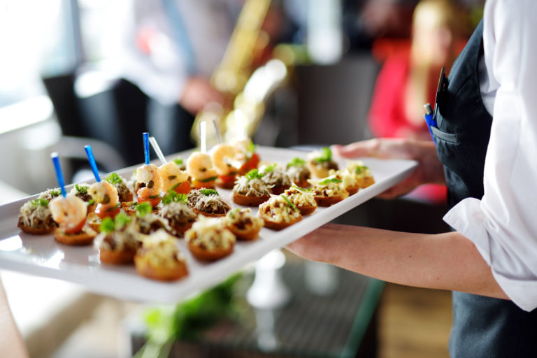 A catering server carries a plate of small meat and seafood appetizers at an event.