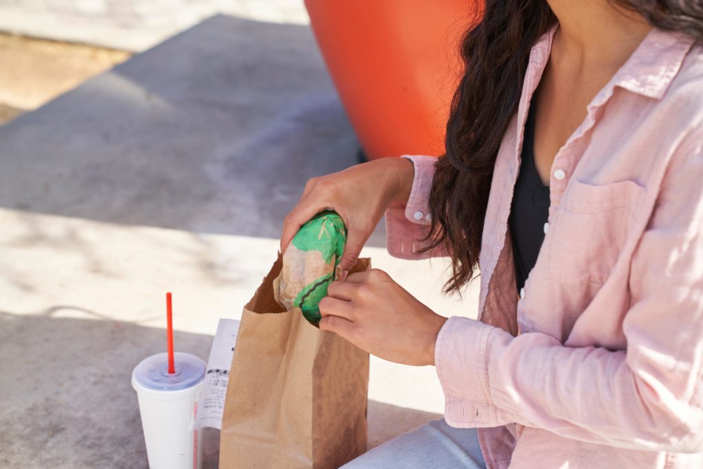 A woman eating fast food outside taking a wrapped burger out of a paper bag.
