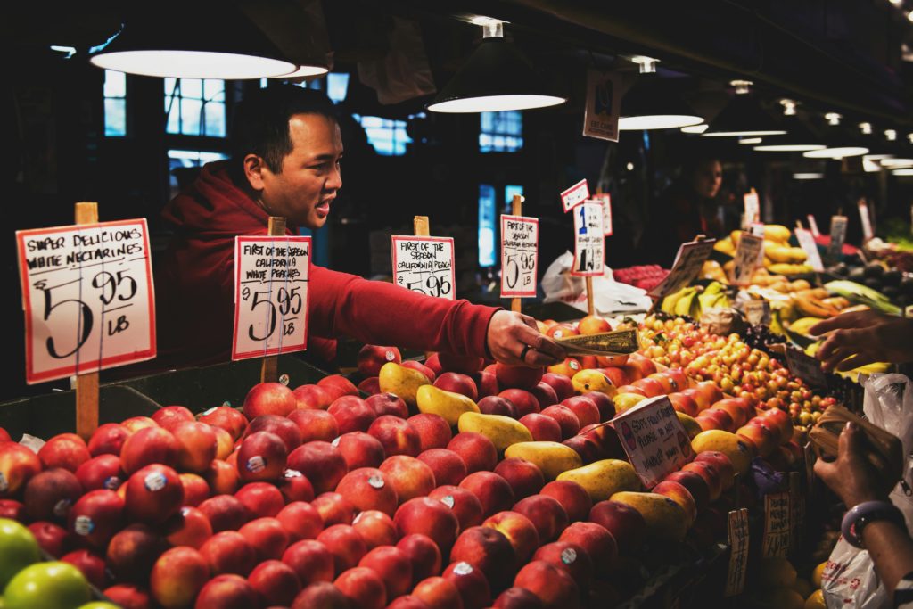 A customer exchanges cash with a farmers market vendor over a display of peaches in an indoor market.