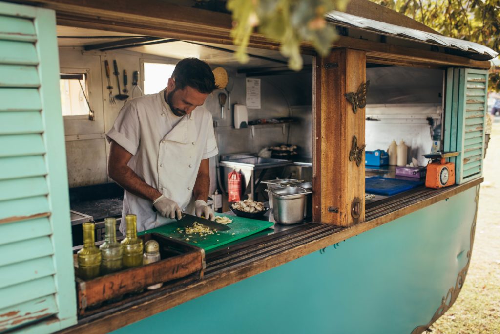 A man in a chef uniform dicing vegetables on a cutting board inside a turquoise food trailer.